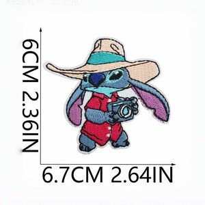 May include: An embroidered patch of a blue character wearing a tan hat with a teal band and a red shirt, holding a camera. The patch measures 2.36 inches in height and 2.64 inches in width as indicated by the black measurement lines and text.
