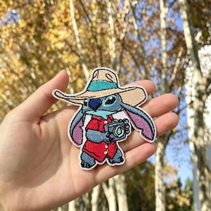 May include: An embroidered patch featuring a blue alien character wearing a tan wide-brimmed hat with a teal band and a red button-down shirt. The character is holding a black camera with a visible lens, and the patch has a white border.