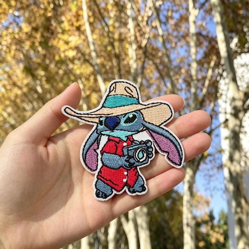 May include: An embroidered patch featuring a blue alien character wearing a tan wide-brimmed hat with a teal band and a red button-down shirt. The character is holding a black camera with a visible lens, and the patch has a white border.