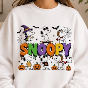 May include: A white crewneck sweatshirt featuring a colorful graphic of Snoopy in various Halloween costumes, including a witch and aviator. The text SNOOPY appears in a dripping, multi-colored font above pumpkins, bats, and spiderwebs.