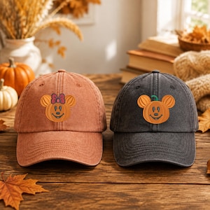 Puede incluir: Dos gorras de béisbol, una en naranja desteñido y otra en gris carbón, cada una con un personaje de calabaza bordado con orejas redondas. La gorra naranja incluye un pequeño lazo rojo en la calabaza, mientras que la gris tiene un tallo verde.