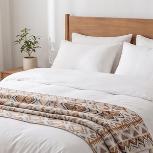 Könnte beinhalten: Ein weißes Bett mit einem hölzernen Kopfteil und einer gewebten Tagesdecke am Fußende. Die Decke zeigt ein sich wiederholendes geometrisches Muster in Beige-, Braun-, Schwarz- und Cremetönen mit einer strukturierten Strickoptik und Fransen.