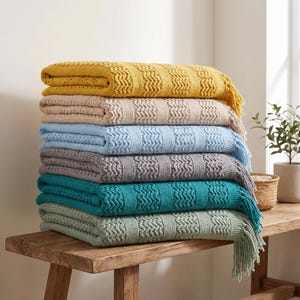 Könnte beinhalten: Ein Stapel aus sechs gefalteten Strickdecken mit strukturiertem Zickzackmuster und Fransen an den Rändern. Die Decken sind in Senfgelb, Beige, Hellblau, Grau, Petrol und Salbeigrün gehalten und ordentlich übereinander gestapelt.