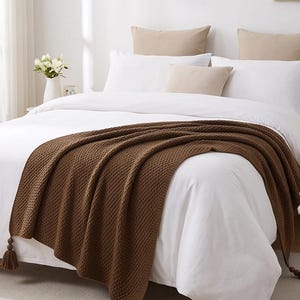 May include: A dark brown knitted throw blanket with a textured waffle pattern and decorative tassels at the corners, draped over a white bedspread. The blanket features a consistent woven design and hangs over the edge of the mattress.