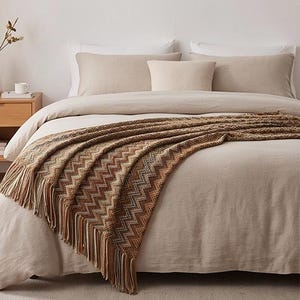 May include: A knitted throw blanket featuring a multi-coloured chevron pattern in shades of brown, tan, and cream. The blanket is draped over a bed with beige bedding and includes long, decorative fringe along the bottom edge of the fabric.