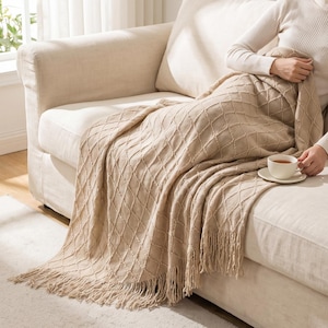 Pode incluir: Uma manta de malha bege com um padrão de diamante em relevo e acabamento com franjas nas bordas. A manta está disposta sobre uma superfície, exibindo a trama texturizada e a cor neutra do tecido em uma visão clara e detalhada.