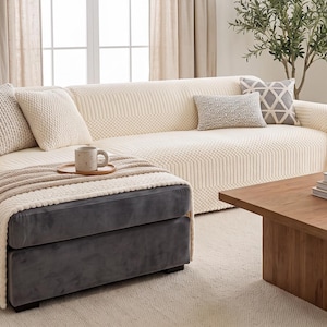 May include: A cream-colored textured sofa cover draped over a sectional sofa with a dark gray velvet ottoman section. The cover features a waffle-knit pattern and is accented with several decorative throw pillows in shades of gray and cream.