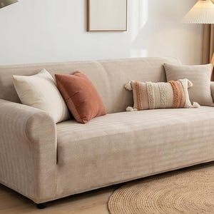 May include: A beige sofa covered in a textured fabric slipcover with rolled arms. The sofa is decorated with two solid beige square pillows, one solid rust-coloured square pillow, and one rectangular lumbar pillow featuring cream and rust stripes.