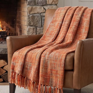 Könnte beinhalten: Eine gewebte Wohndecke mit einem strukturierten Muster in Orange-, Rost- und Cremetönen. Die rechteckige Decke hat eine dicke, grobe Strickstruktur und ist am unteren Rand mit langen, geknoteten Fransen versehen.