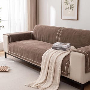 May include: A brown sofa cover with a textured surface and white piping trim, draped over a cream-colored sofa. Two stacked books with the text DEFION and DESIGN are placed on the seat, along with a cream-colored knitted throw blanket.