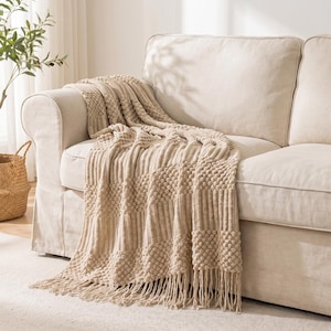 May include: A beige throw blanket featuring a textured pattern of alternating vertical ribbed stripes and raised bobble knit sections. The blanket is draped over a sofa and finished with long, knotted fringe tassels along the bottom edge.
