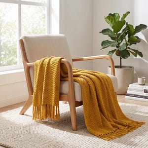 May include: An armchair with light beige upholstery and a wooden frame, draped with a mustard yellow textured knit throw blanket featuring fringe edges. The blanket covers the seat and backrest, extending down to the floor on one side.
