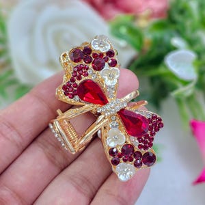May include: A gold-toned metal hair claw clip shaped like a butterfly, featuring large red teardrop-shaped crystals, smaller round deep red rhinestones, and white floral accents. The clip has a spring mechanism and clear crystals along the centre.