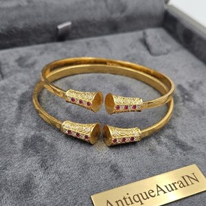 May include: A pair of gold-coloured bangles with intricate patterns and red and white gemstones. The open-ended bangles have decorative flared ends. The jewellery is presented in a grey velvet-lined box. A gold plaque reads "AntiqueAuraIN".