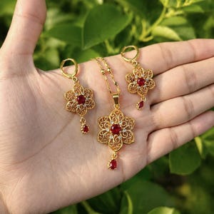 Puede incluir: Un conjunto de joyas de color dorado con un diseño floral y piedras preciosas rojas. El conjunto incluye un colgante de collar, pendientes y un colgante a juego. La imagen también incluye el texto "AntiqueAuralN" y "Timeless Beauty, Just for you."