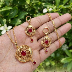 Puede incluir: Juego de joyas doradas con collar y pendientes. El collar tiene un colgante con una piedra roja central y una lágrima a juego. Los pendientes tienen un diseño similar. El conjunto se muestra sobre un fondo verde.