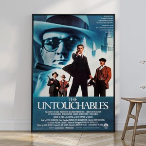 Puede incluir: Un póster de película enmarcado de The Untouchables con un gran retrato en tonos azules de un hombre con sombrero fedora y gafas, y tres hombres frente a un horizonte urbano. El título está impreso en blanco en la parte inferior con créditos.