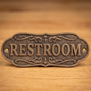 Könnte beinhalten: Bronzefarbenes Toilettenschild mit verzierten Schnörkeln und dem Wort "RESTROOM" in fetten Buchstaben. Das Schild hat einen strukturierten Hintergrund und ist auf einer Holzoberfläche montiert. Dekorativ und funktional.