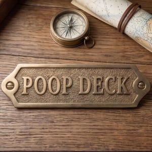 May include: A brass-colored metal sign with the words "POOP DECK" in bold, raised lettering. The sign is rectangular with decorative edges and is mounted with screws. A vintage compass and rolled map are in the background.