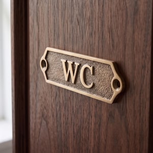 May include: A rectangular brass sign with a textured bronze-colored background and raised gold-toned letters reading WC. The sign features rounded ends with two circular screw holes for mounting and is attached to a dark brown wood surface.