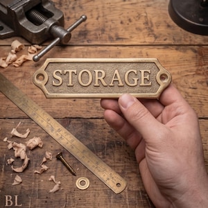 Könnte beinhalten: Ein rechteckiges Messingschild mit abgeschrägten Kanten und strukturiertem Hintergrund, das das Wort STORAGE in erhabenen Großbuchstaben zeigt. Das Schild verfügt über zwei kreisförmige Befestigungslöcher an den Enden und polierte Buchstaben.