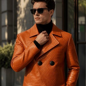 May include: A double-breasted jacket in a vibrant orange hue featuring a textured crocodile-embossed pattern. The garment includes wide lapels, two large black circular buttons, and long sleeves, worn over a black high-neck sweater and sunglasses.