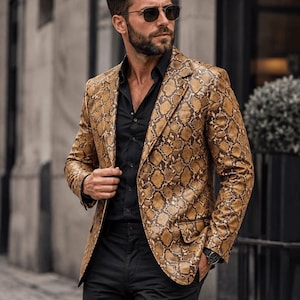 Könnte beinhalten: Ein beigefarbener Blazer mit braunem Schlangenledermuster, fallendem Revers, zwei Vordertaschen mit Patte und einem einreihigen Knopfverschluss. Die Jacke wird über einem einfarbigen schwarzen Hemd mit schwarzer Hose getragen und hat eine Passform.