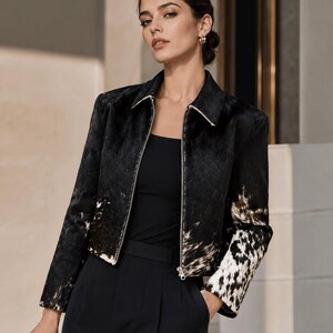 May include: A black and white cowhide jacket with a zipper closure. The jacket has a collar and long sleeves. The model is wearing a black top and black pants. The jacket has a white and brown spotted pattern on the sleeves.
