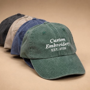 Może przedstawiać: Rząd czapek bejsbolowych w różnych kolorach: czarnym, beżowym, niebieskim i zielonym. Zielona czapka ma biały haft "Custom Embroidery EST. 2026". Czapki wykonane są z miękkiego, teksturowanego materiału.