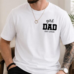 May include: A white short-sleeved t-shirt featuring black text on the upper left chest area. The text is arranged in three lines and reads "girl" in a cursive script, "DAD" in a bold block font, and "EST.2026" in a smaller, simple font.
