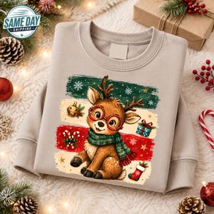 Può includere: Una felpa beige a girocollo con la grafica di una renna marrone che indossa occhiali rossi e una sciarpa verde. Il design presenta strisce orizzontali verdi, crema e rosse, decorate con un campanellino dorato, un pacchetto regalo e una calza.