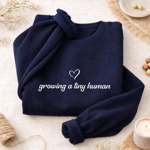 Può includere: Una felpa girocollo blu navy con un'icona a forma di cuore bianco e la scritta bianca "growing a tiny human" stampata al centro del petto. Il capo presenta maniche lunghe con polsini a coste e uno scollo a coste.