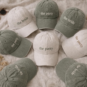 Puede incluir: Una colección de gorras de béisbol en tonos verde oliva lavado y blanco roto. Las gorras presentan texto bordado en los paneles frontales, algunas con la frase the bride y otras the party en una fuente sencilla sin remates.