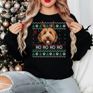 Può includere: Una felpa nera a maniche lunghe con la grafica di un cane marrone con corna di renna. Il design include motivi bianchi, rossi e verdi di impronte di zampe e alberi di pino, con il testo HO HO HO HO stampato in bianco al centro.