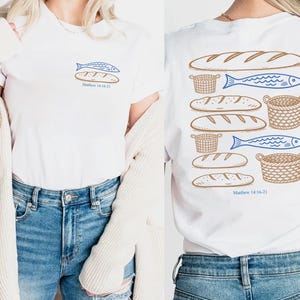 May include: A white t-shirt featuring a small graphic on the front chest of a blue fish and a brown loaf of bread with the text Matthew 14:16-21. The back displays a larger graphic of multiple brown loaves of bread, blue fish, and woven baskets.