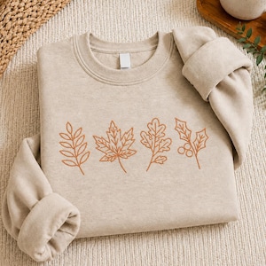 Könnte beinhalten: Ein beigefarbenes Sweatshirt mit Rundhalsausschnitt und einer Stickerei aus vier verschiedenen botanischen Elementen in rostorangefarbenem Garn. Das Motiv zeigt ein farnartiges Blatt, ein Ahornblatt, ein Eichenblatt und einen Zweig mit Beeren.