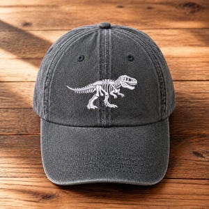 May include: A charcoal grey baseball cap featuring a white embroidered dinosaur skeleton graphic on the front panel. The cap has a six-panel construction with a curved brim, stitched eyelets, and a washed fabric texture throughout the design.