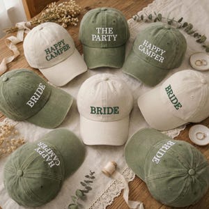 May include: An assortment of baseball caps in cream and olive green. Each cap has embroidered text, including "HAPPY CAMPER", "THE PARTY", and "BRIDE". The caps are displayed on a wooden surface, with decorative elements.