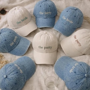 May include: A collection of baseball caps in denim blue and off-white fabric. Some caps feature the embroidered text "the bride" in green or white, while others display the embroidered text "the party" in dark brown or white thread.