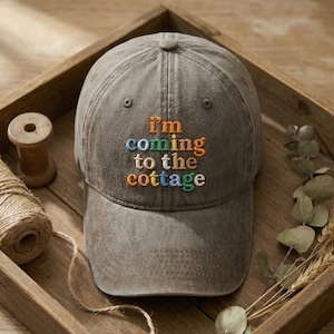 May include: A gray baseball cap with the embroidered phrase "I'm coming to the cottage" in colorful letters. The hat is made of a soft fabric and has a curved brim. The cap is sitting in a wooden tray.