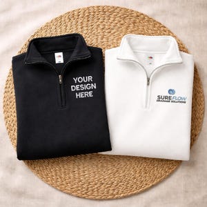 Op de afbeelding: Een zwart sweatshirt met een kwartrits en de tekst YOUR DESIGN HERE en een wit sweatshirt met een kwartrits en de tekst SUREFLOW DRAINAGE SOLUTIONS. Beide kledingstukken zijn voorzien van een zilverkleurige rits en een hoge kraag.