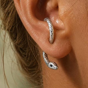 May include: A silver-toned ear cuff shaped like a snake that wraps around the ear cartilage. The metal features a textured, scale-like pattern throughout its body, and the snake head is detailed with a small, dark, circular eye accent.