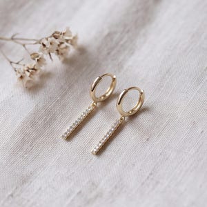 May include: A pair of gold hoop earrings featuring a dangling bar charm. Each bar is encrusted with a row of small, clear, sparkling stones. The earrings have a polished finish and a hinged closure mechanism for secure wear.