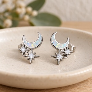 Pode incluir: Um par de brincos de tarracha prateados em forma de meia-lua incrustados com opala branca iridescente e dois pequenos detalhes em forma de estrela com pedras transparentes brilhantes e acabamento em metal polido.