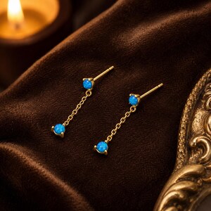 May include: A pair of gold-toned stud earrings featuring a post back, a small round blue opal stone at the top, and a delicate gold chain dangling down to a second matching round blue opal stone at the bottom of each earring.