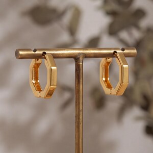 May include: A pair of gold-toned hoop earrings with a distinct hexagonal shape. The earrings feature a smooth, polished metallic finish and are displayed hanging from a horizontal metal bar stand, showcasing their geometric design and structure.