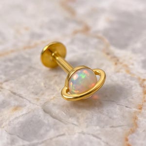 May include: A gold-toned labret stud body jewelry piece featuring a round, iridescent white opal cabochon set in the center of a thin, circular gold ring, resembling a planet with a ring, with a flat circular back disc.