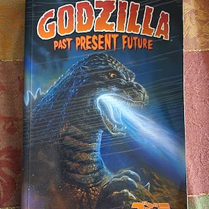Könnte beinhalten: Ein Buchcover mit einer Godzilla-Illustration und dem Titel "Godzilla Past Present Future" in orangefarbenem Text. Die Godzilla-Illustration ist eine Nahaufnahme des Kopfes und Halses des Monsters, mit offenem Maul und einem blauen Energie-Strahl, der aus seinem Maul schießt. Der Hintergrund ist dunkelblau mit einem Sternenhimmel. Das japanische Wort für Godzilla, "ゴジラ", ist in orange unten auf dem Cover gedruckt.
