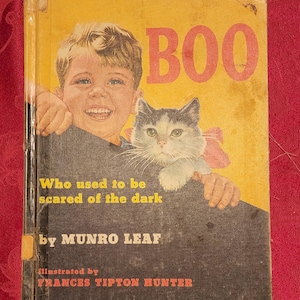 以下が含まれることがあります： 黄色の本の表紙に、赤い文字で「BOO」というタイトルが書かれている。表紙には、本の端から顔を出している少年と猫が描かれている。タイトルの下には、「暗闇を恐れていた人」と「MUNRO LEAF 著」という文章が書かれている。この本は、Frances Tipton Hunterによってイラストが描かれている。