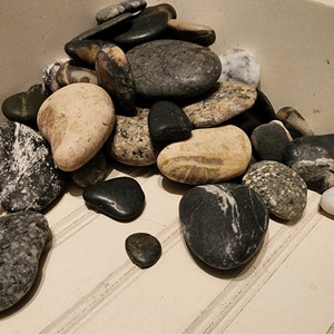 May include: A pile of smooth, gray, black, and white stones in a white box. The stones are various sizes and shapes.